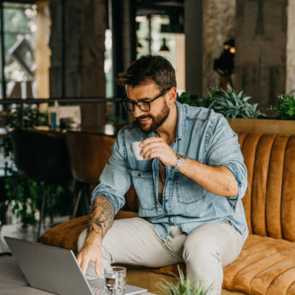Spontaneous image of a handsome looking bearded, tattooed male, owner of a small company, working on his laptop while sitting in a launch of a hotel, enjoying his coffee. He's wearing eyeglasses on, urban stylishly dressed, wearing a denim shirt with a cream pants, typing with a soft smile on his face.